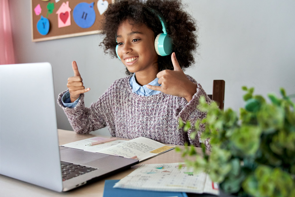 a girl using hand signs with headphones on and smiling while communicating with someone in a laptop